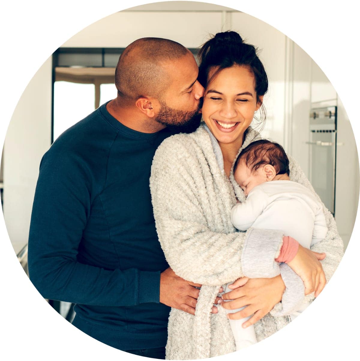 A woman holding a newborn baby in a kitchen, her husband is behind her kissing her cheek